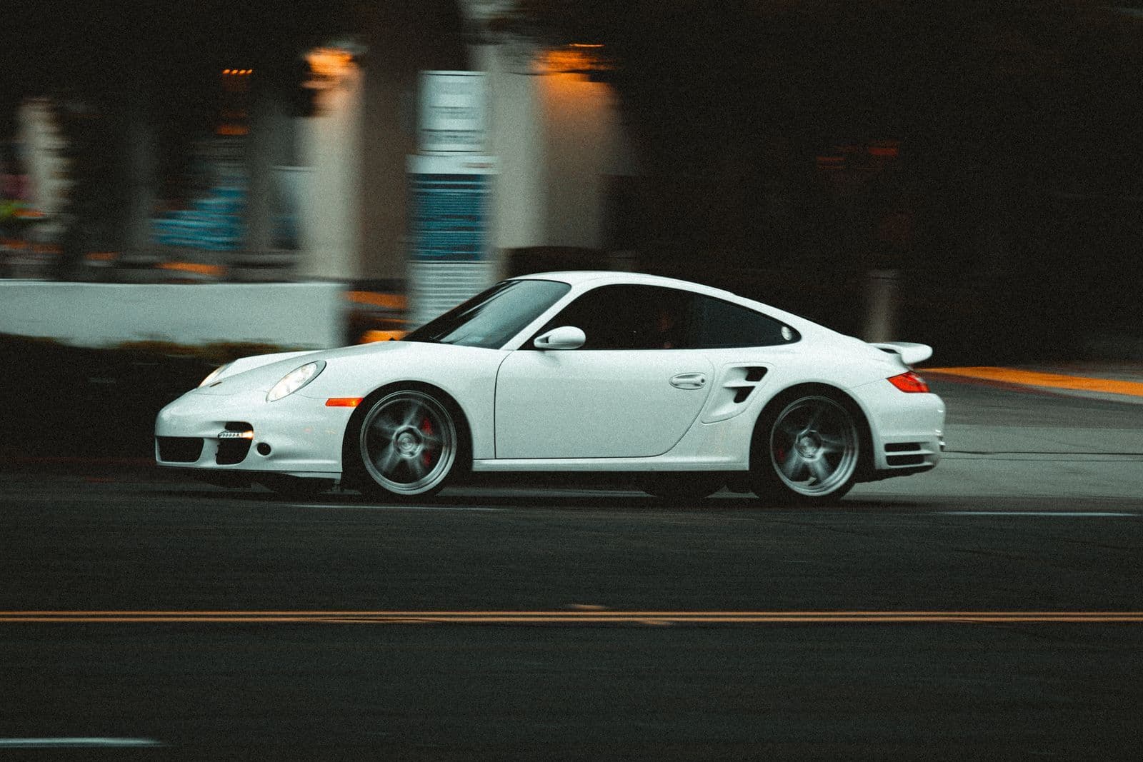 A white Porsche 911 sports car photographed in profile as it moves along a street at dusk.