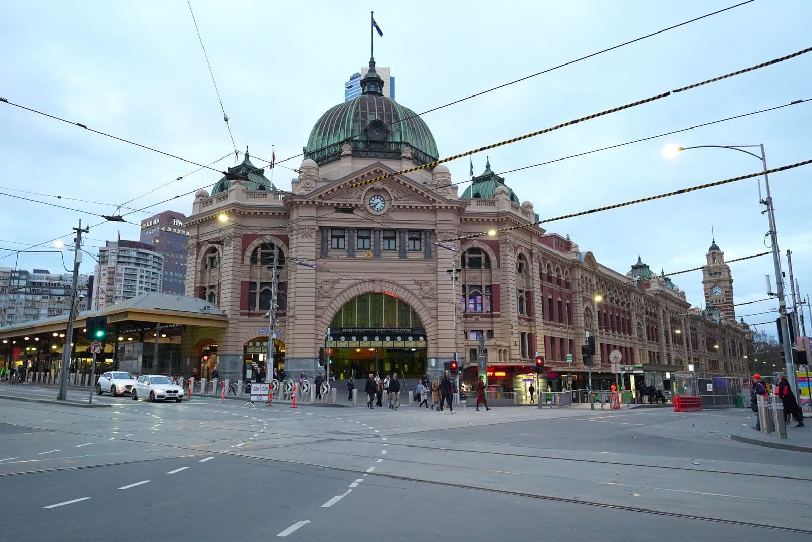 Cars on the road in front of Flinders Street Station in Melbourne at dusk, with tram wires overhead.