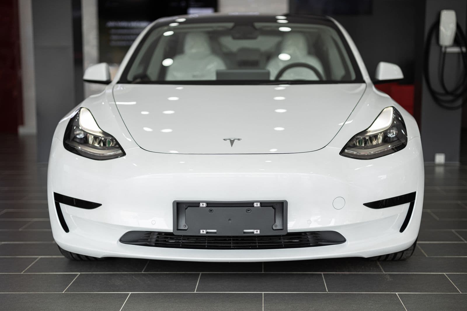 A white Tesla electric sedan photographed head-on inside a showroom, its empty number-plate holder blank and a wall-mounted charger behind it.