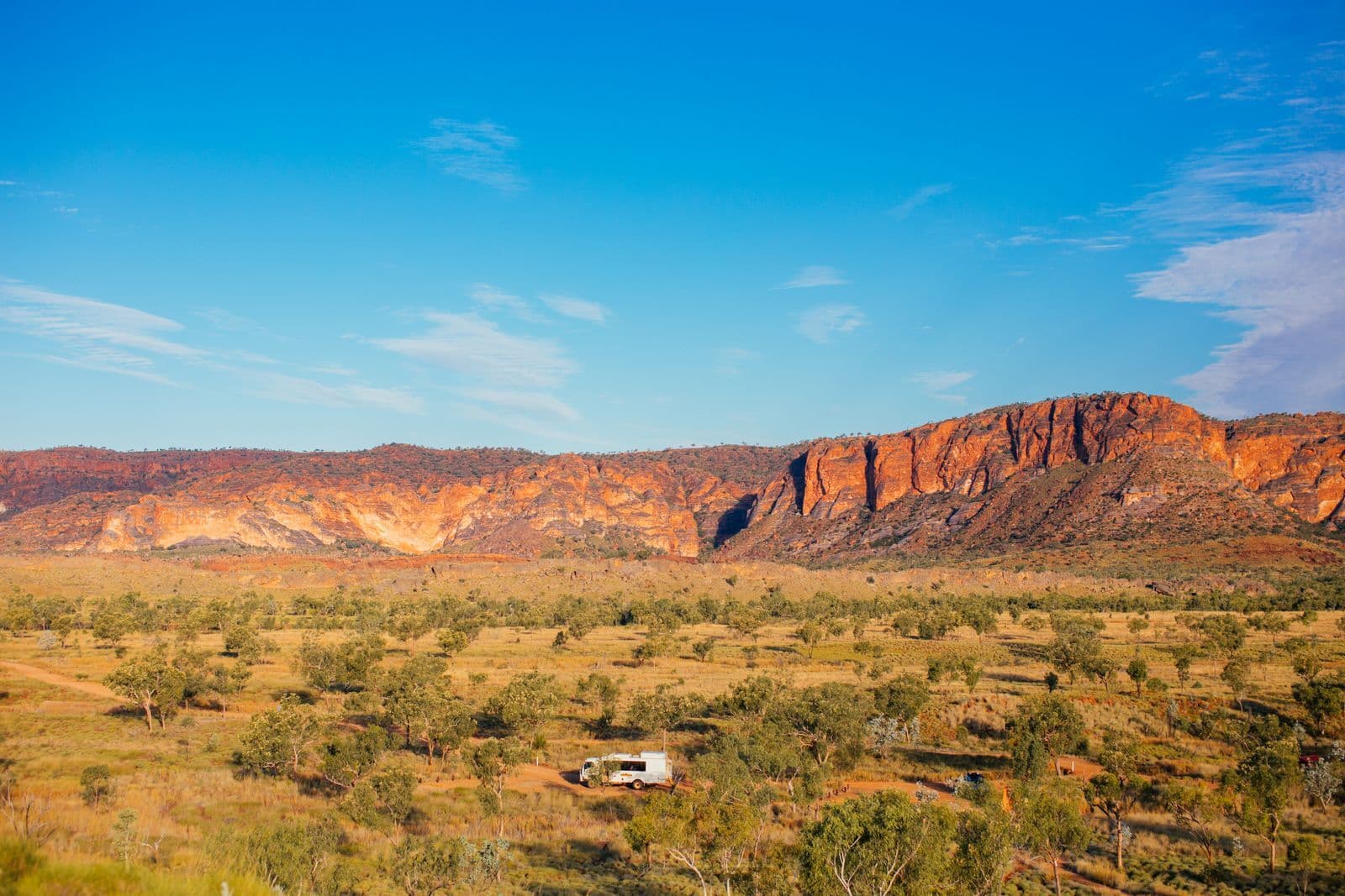 A white four-wheel drive parked among scrub on an outback plain beneath red sandstone cliffs and a blue sky.