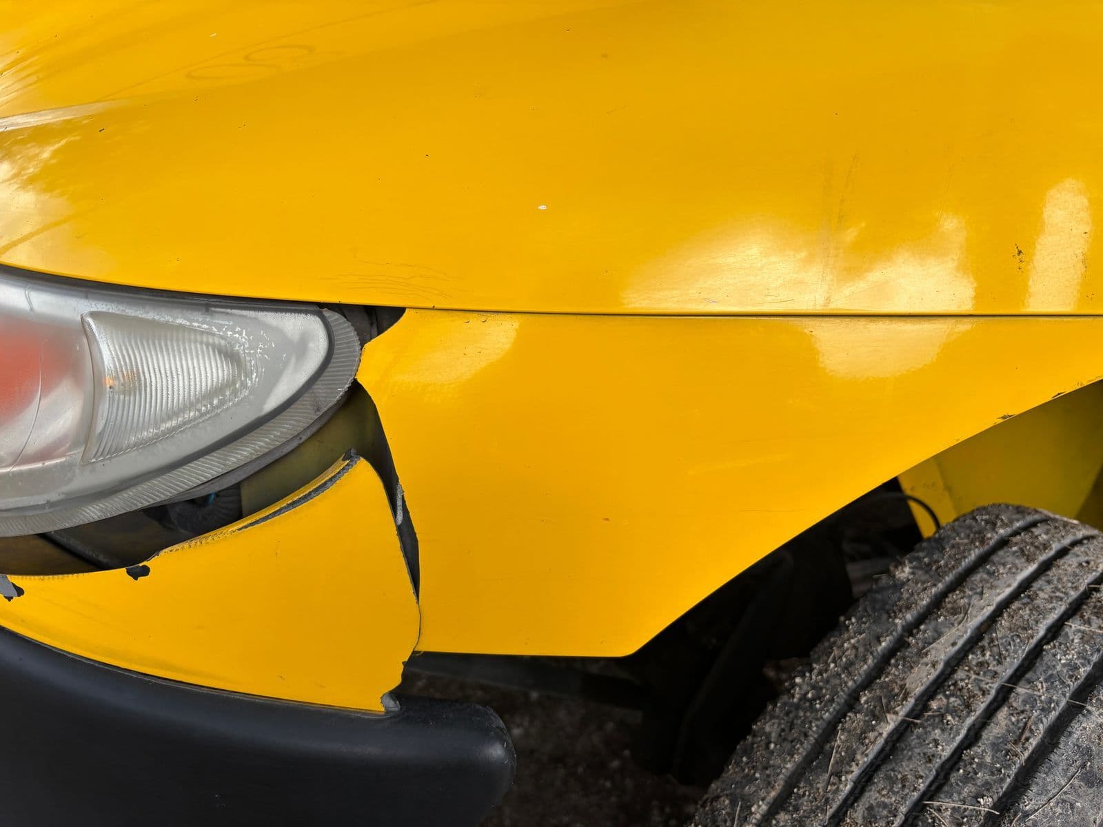 A close-up of a yellow car's cracked front bumper and dented panel beside the headlight.