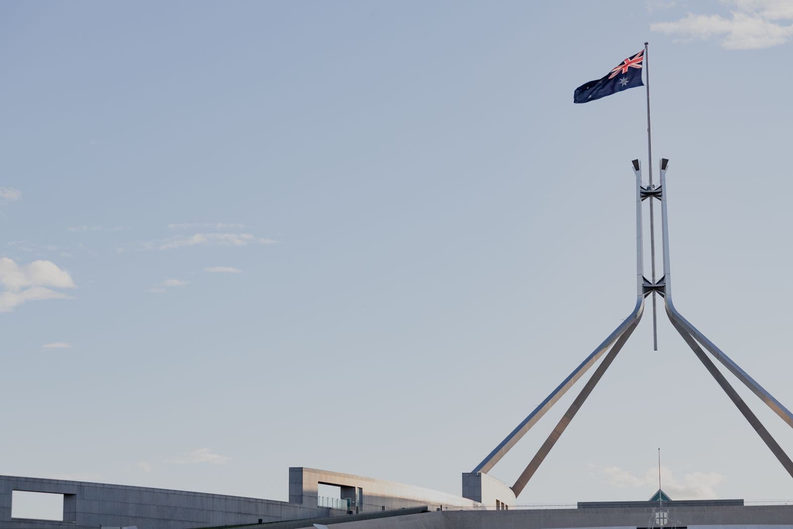 The Australian flag flying above the steel flagpole of Parliament House in Canberra against a pale blue sky.