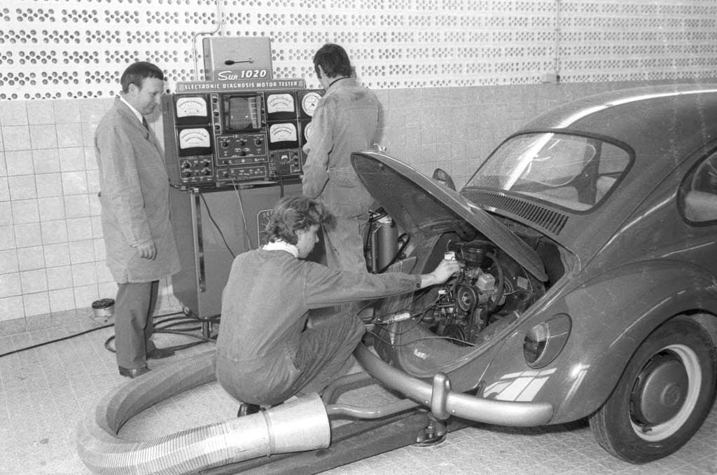 Black-and-white photo of two young apprentices in overalls working on a Volkswagen Beetle's engine beside an electronic engine tester while an older man in a coat watches.
