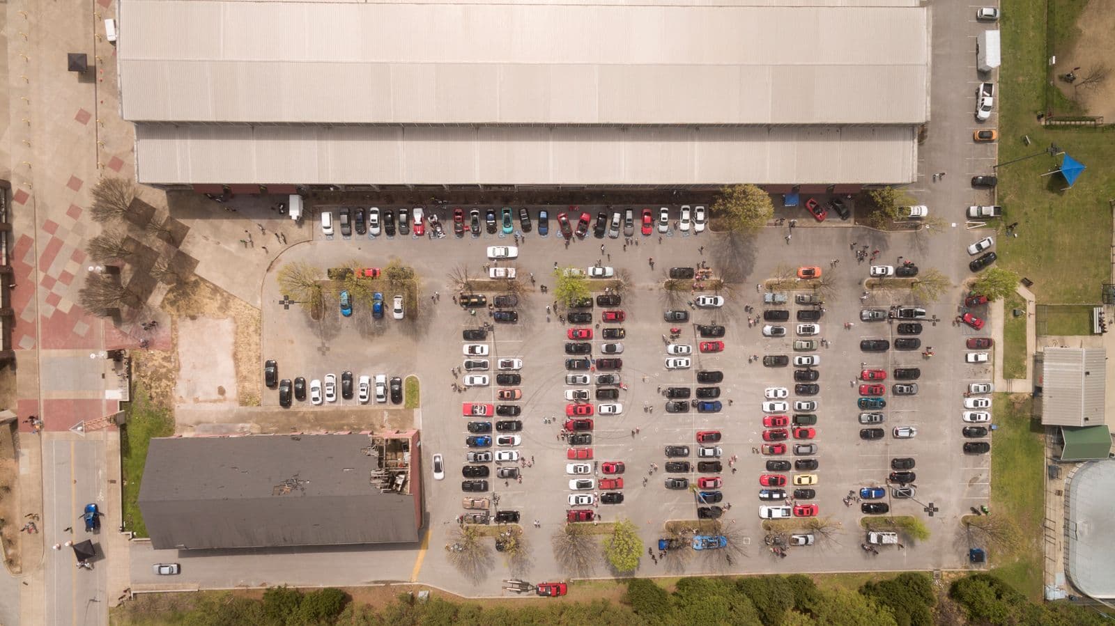 An aerial view of a busy outdoor car park full of parked cars beside a large warehouse-style building.