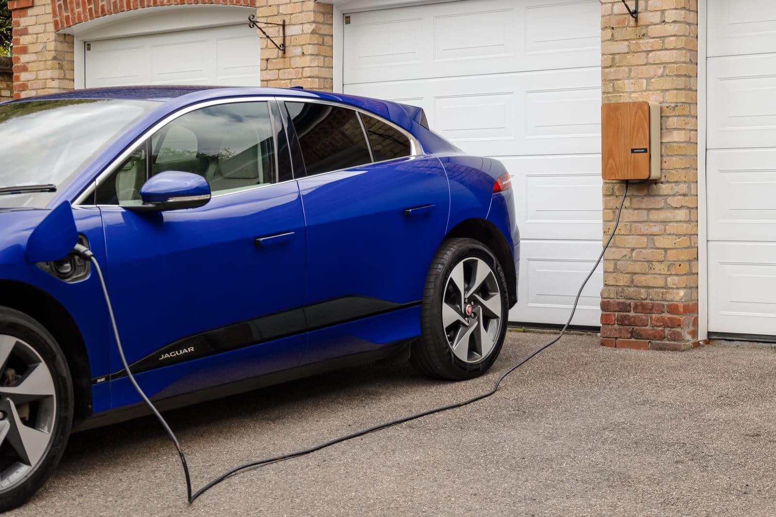 A blue electric SUV parked in a driveway, plugged by a cable into a wood-panelled charger mounted on a brick wall between garage doors.