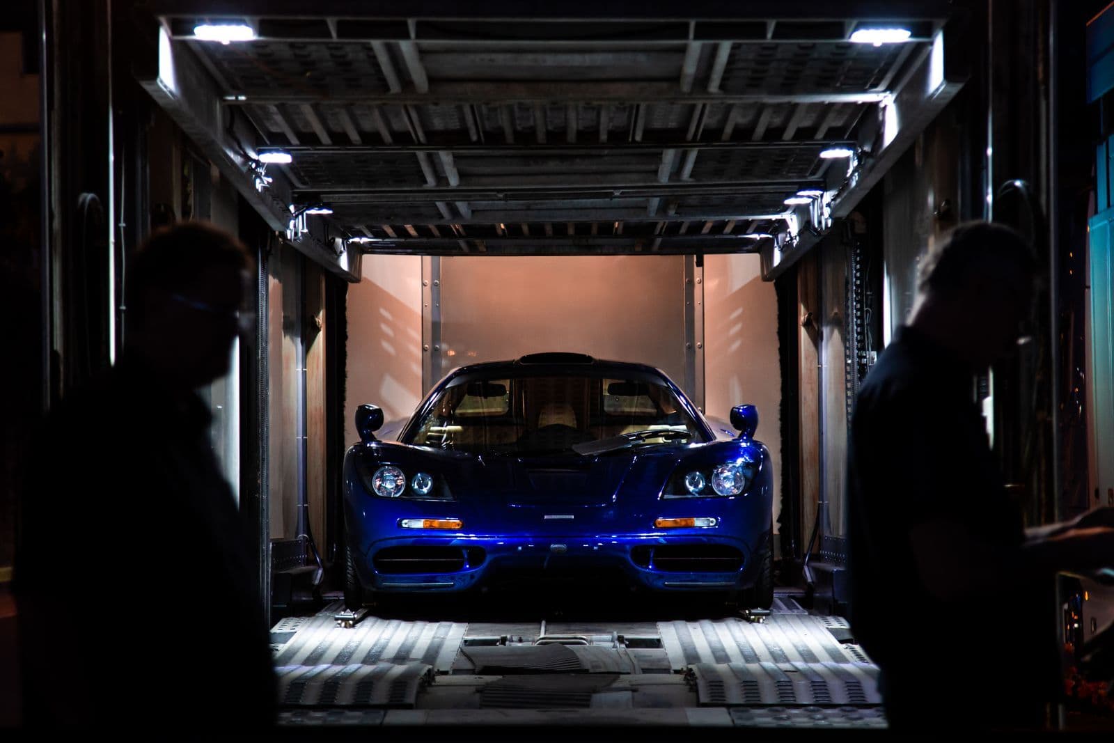 A blue supercar strapped inside the lit interior of a covered car transporter at night, with two people silhouetted in the foreground.