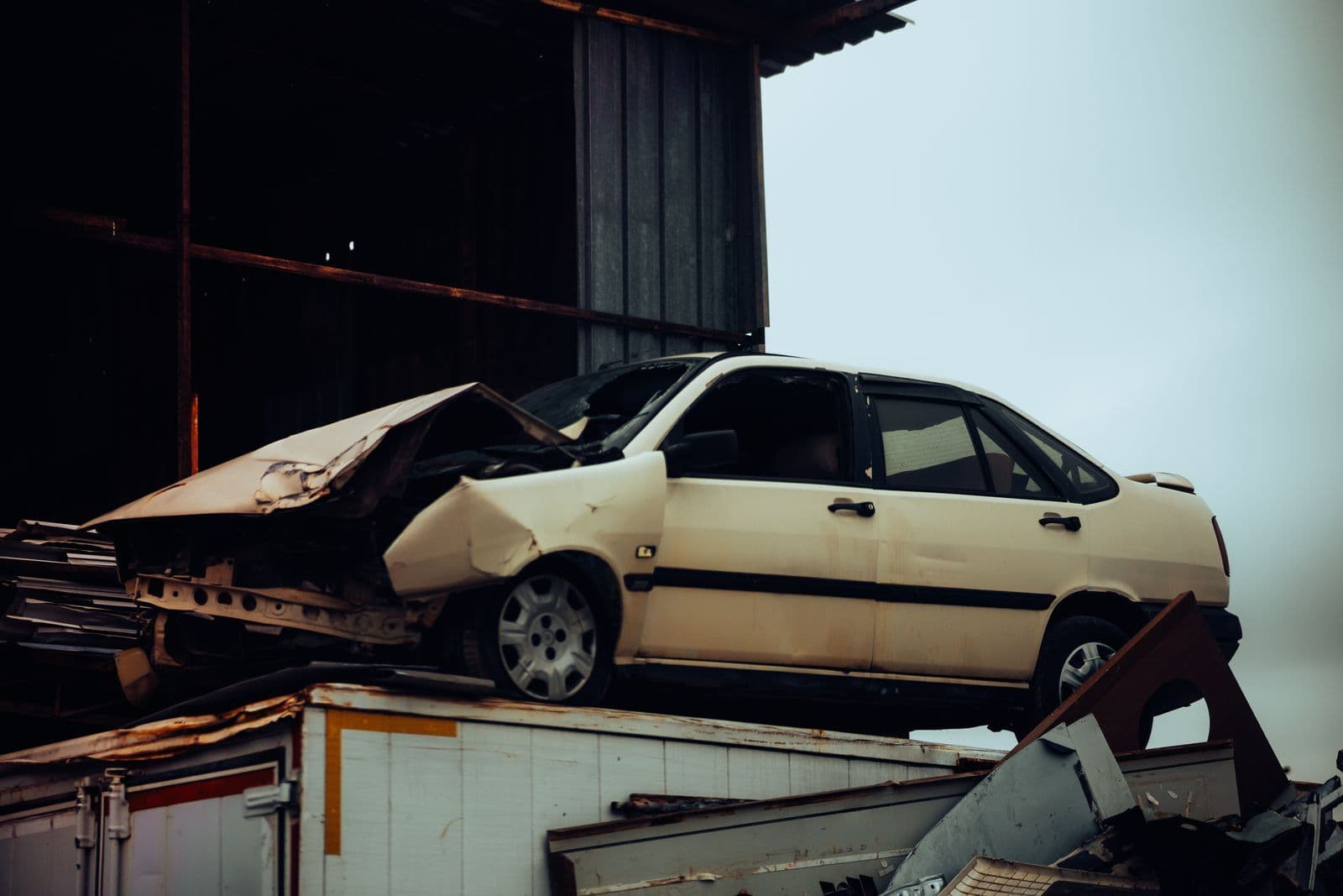 A white 1990s sedan with its front end crushed, resting on a pile of scrap metal beside a shed.