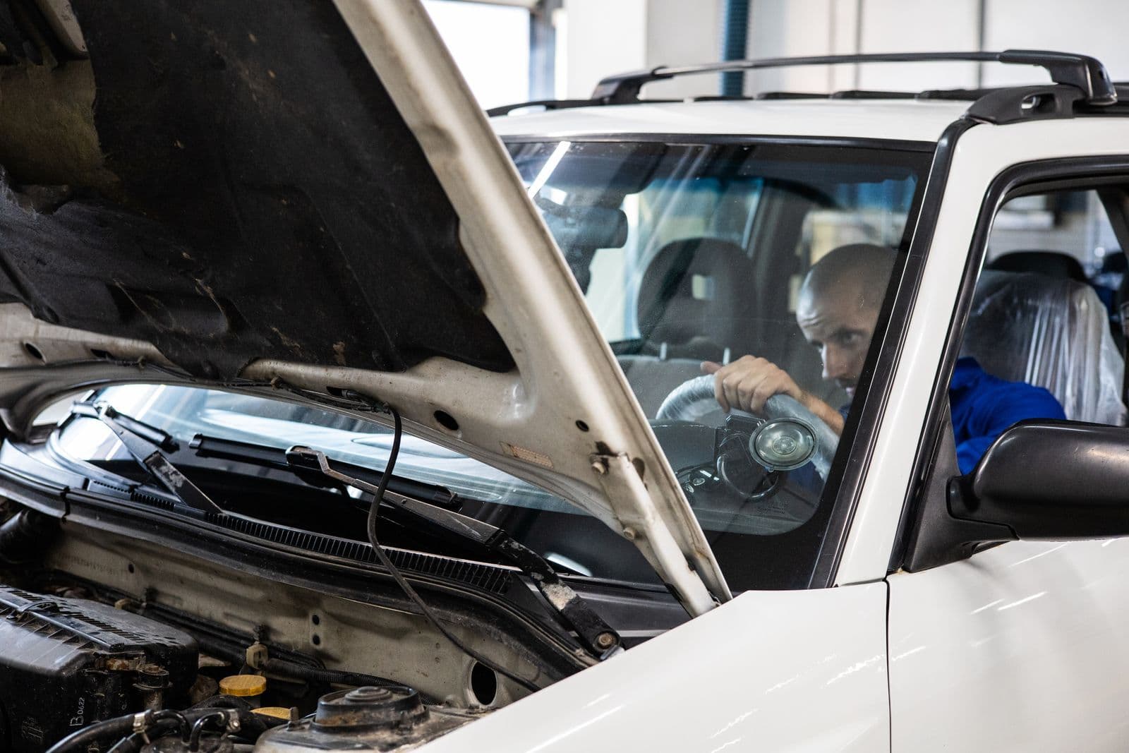 A mechanic sits at the wheel of a white SUV with its bonnet raised inside a workshop.