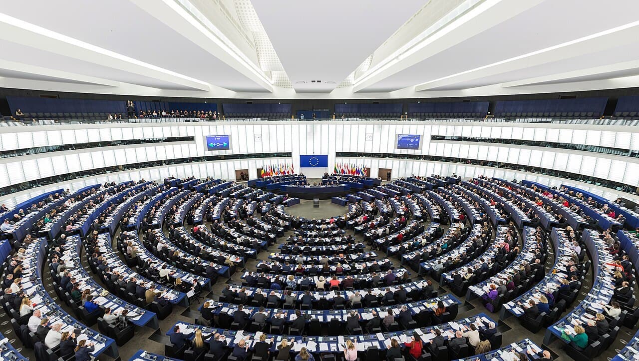 The European Parliament's semicircular debating chamber in Strasbourg, with members seated in rows of blue seats during a session.