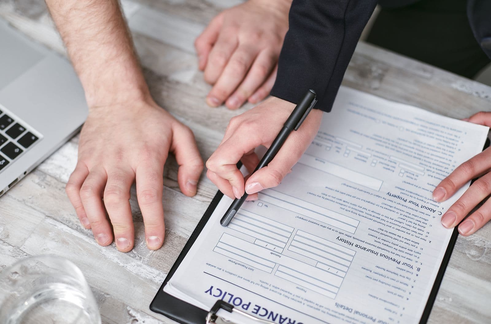 Two people at a desk reviewing and signing a printed insurance policy form on a clipboard, one holding a pen over the page.