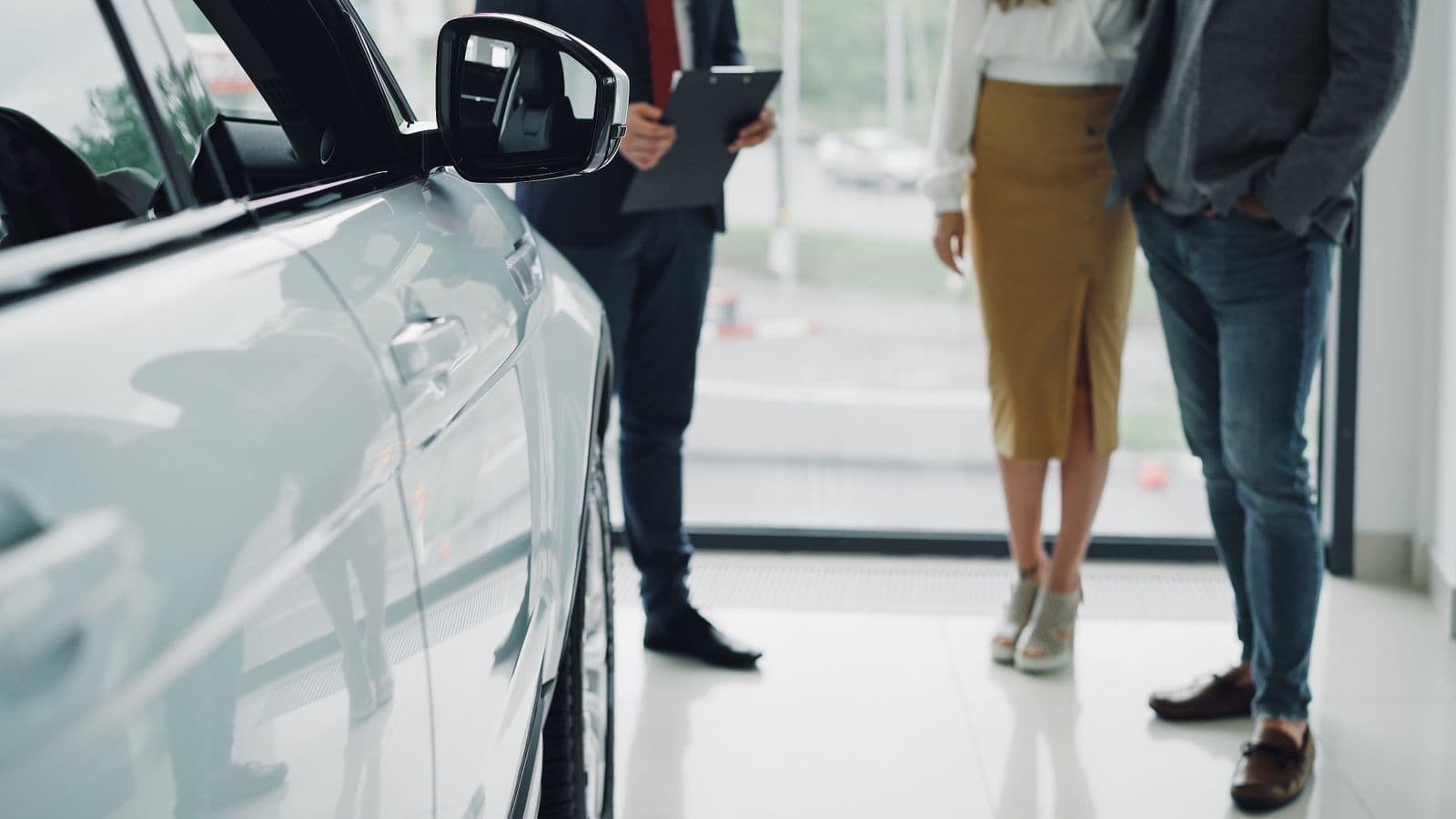 A salesperson holding a clipboard stands with two customers beside a silver car on a bright showroom floor, seen from the shoulders down.