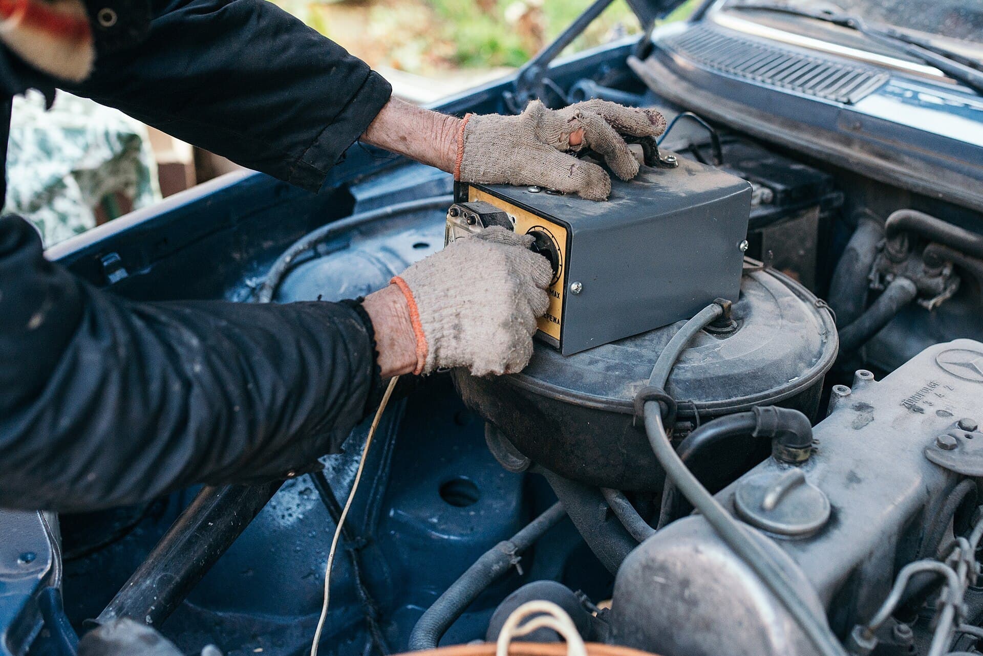 Gloved hands hold a battery tester on top of the open engine bay of an older Mercedes-Benz sedan.