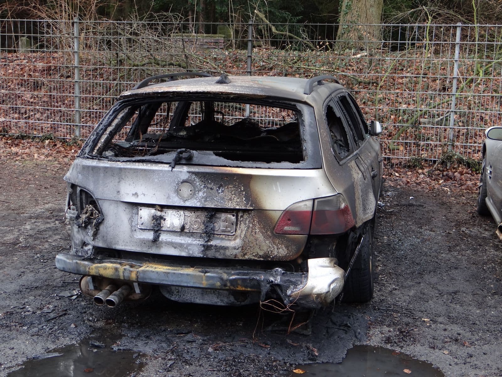 A burnt-out station wagon with scorched panels and blown-out windows standing on gravel beside a wire fence.