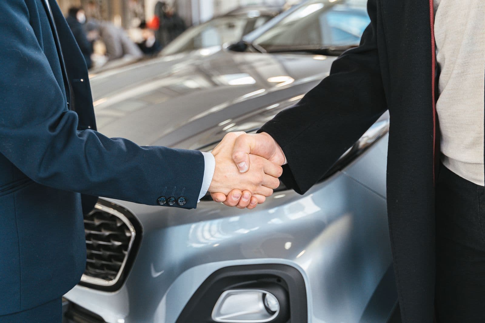 Two people shake hands in front of the bonnet of a silver car, shown from the chest down.