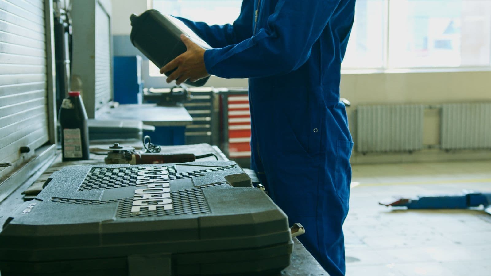 A mechanic in blue overalls holds a black bottle of engine oil beside an open tool case in a workshop.