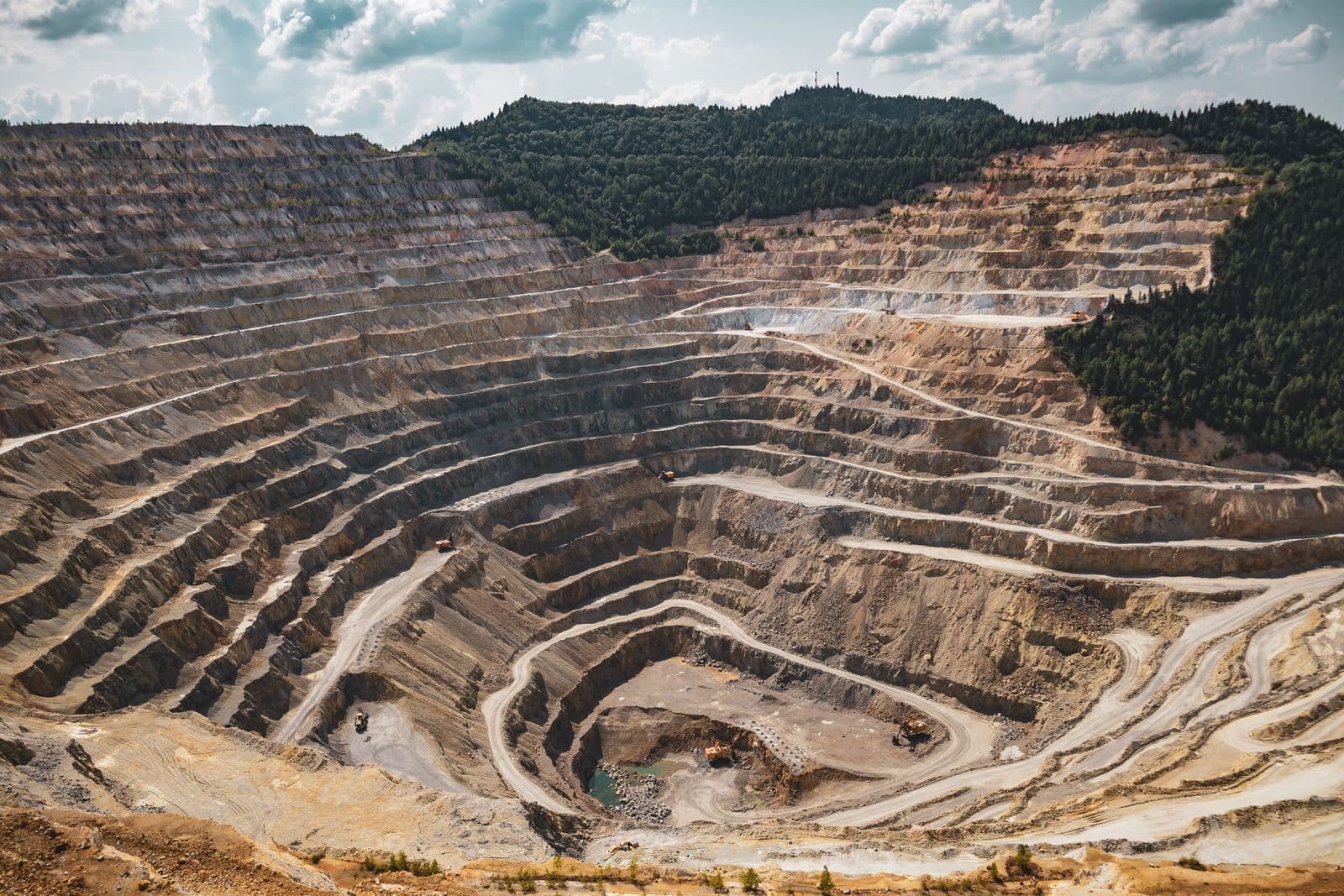 A vast terraced open-pit mine cut into a forested hillside, with haul roads spiralling down to machinery at the bottom.
