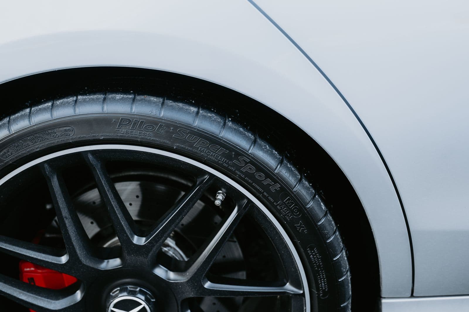 Close-up of a low-profile performance tyre on a black alloy wheel with a red brake caliper, beaded with rain on a silver car.