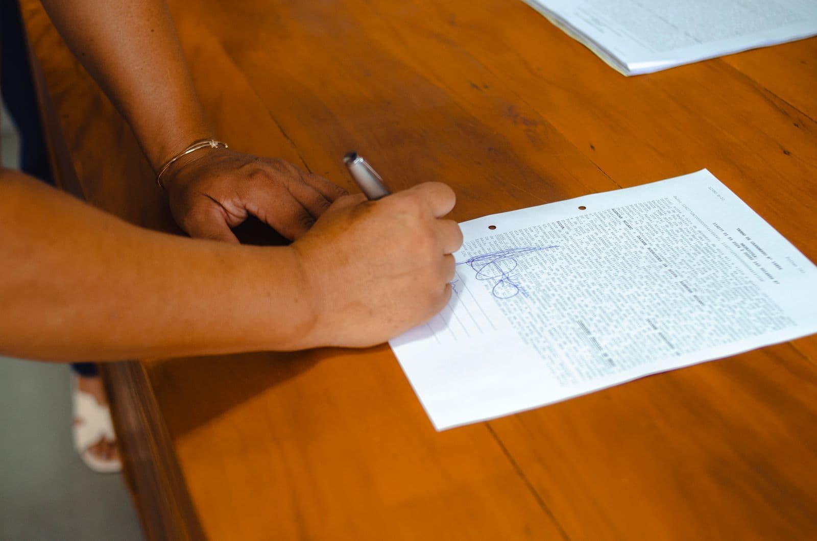 Hands signing a printed form with a pen on a wooden table.