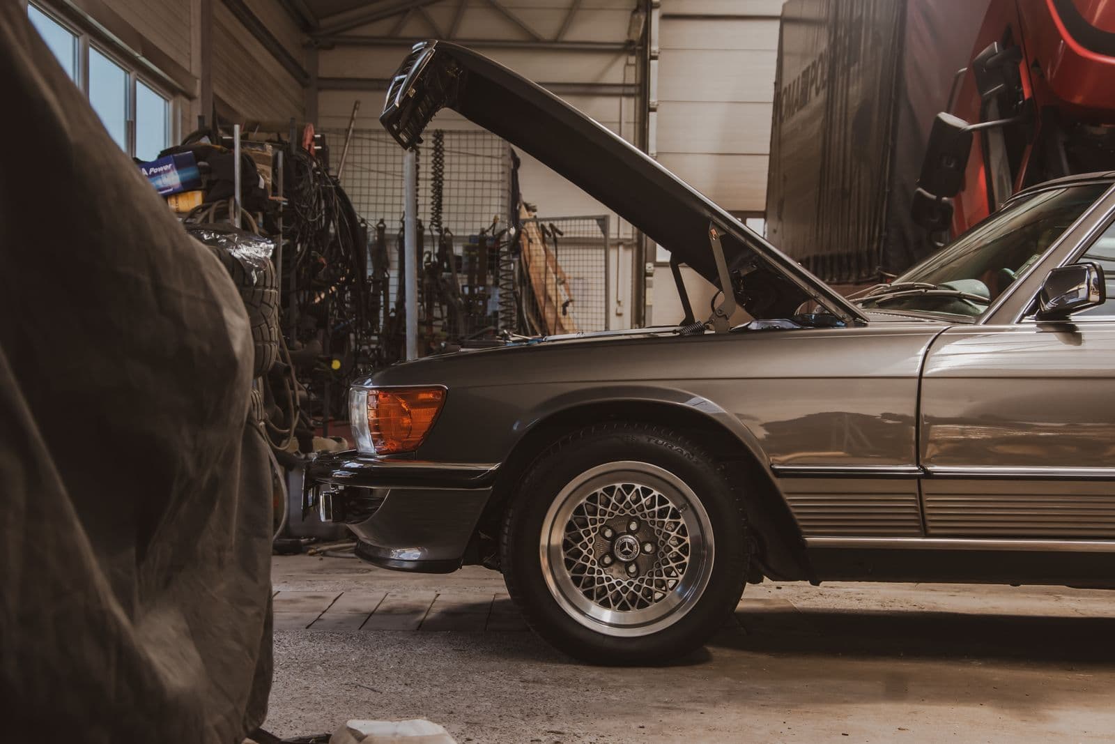 An older grey coupe standing in a workshop with its bonnet propped open, tools and parts racked on the wall behind it.
