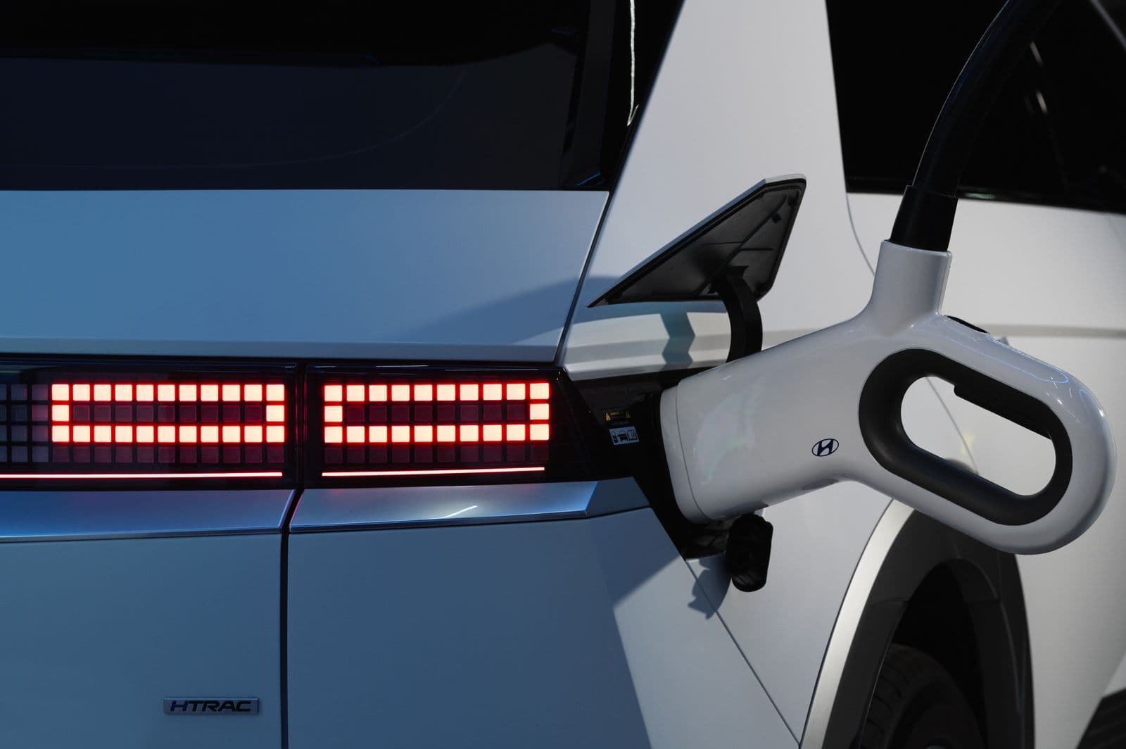 A white charging handle plugged into the side of a white Hyundai Ioniq 5, beside its glowing pixel-style tail lights.