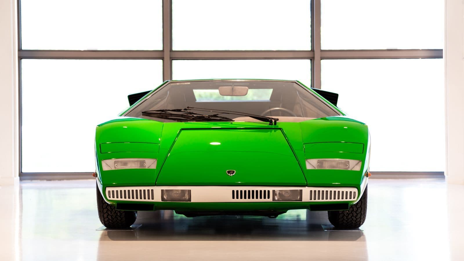 A bright green Lamborghini Countach photographed head-on inside a showroom, its wedge nose and pop-up headlight covers lit by a wall of windows behind.