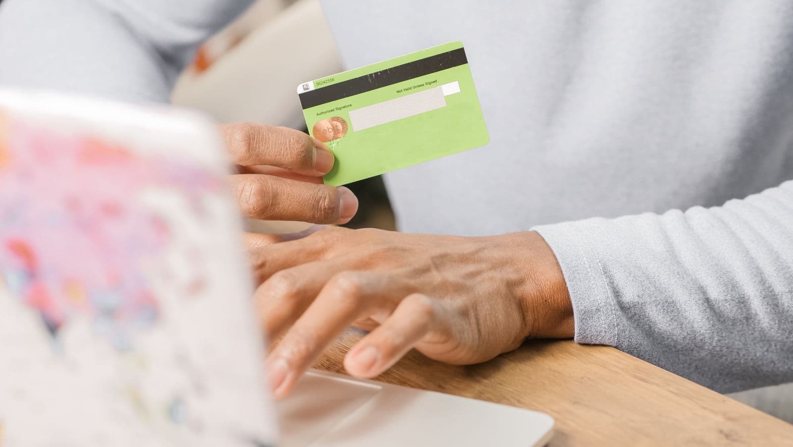 A person holds a green bank card in one hand while typing on a laptop keyboard at a wooden table.