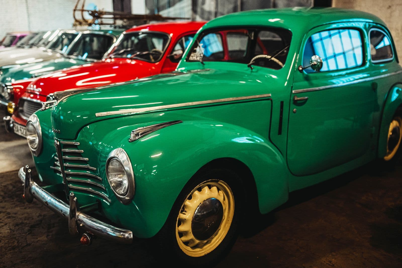 A row of brightly coloured vintage cars parked nose-first inside a workshop, a green one closest to the camera.