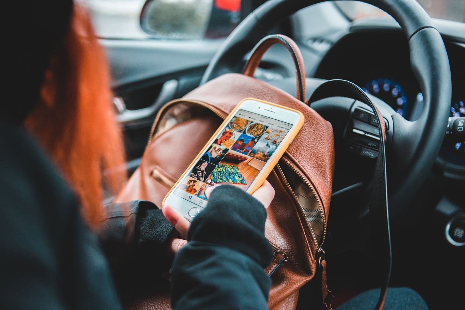 Seen over the shoulder, a person sitting in a car scrolls through a grid of photos on a smartphone, a leather bag on their lap.