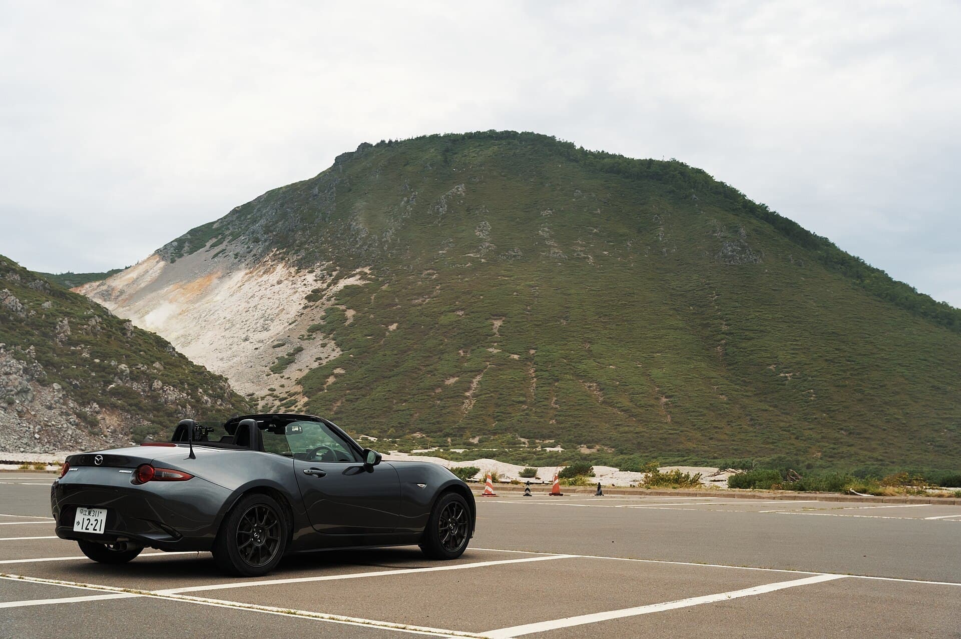 A dark grey Mazda MX-5 roadster with its roof down, parked in an empty car park beneath a green volcanic hill.