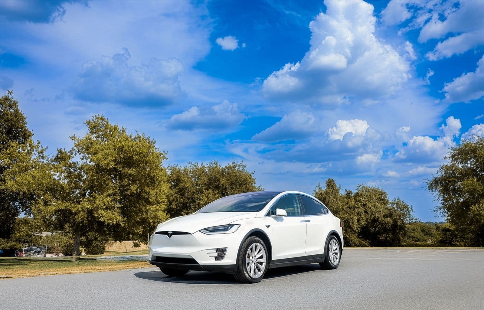 A white electric SUV parked on a quiet country road lined with trees under a bright blue sky with scattered cloud.