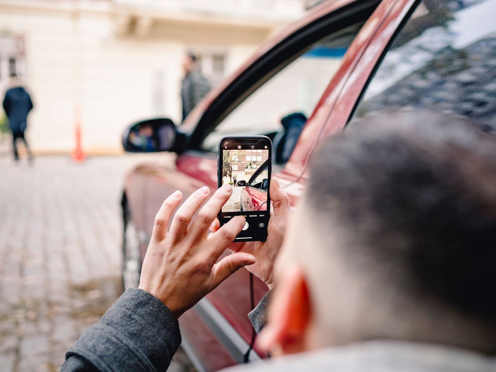 A man seen from behind photographs the side of a red car with his smartphone on a cobbled street.