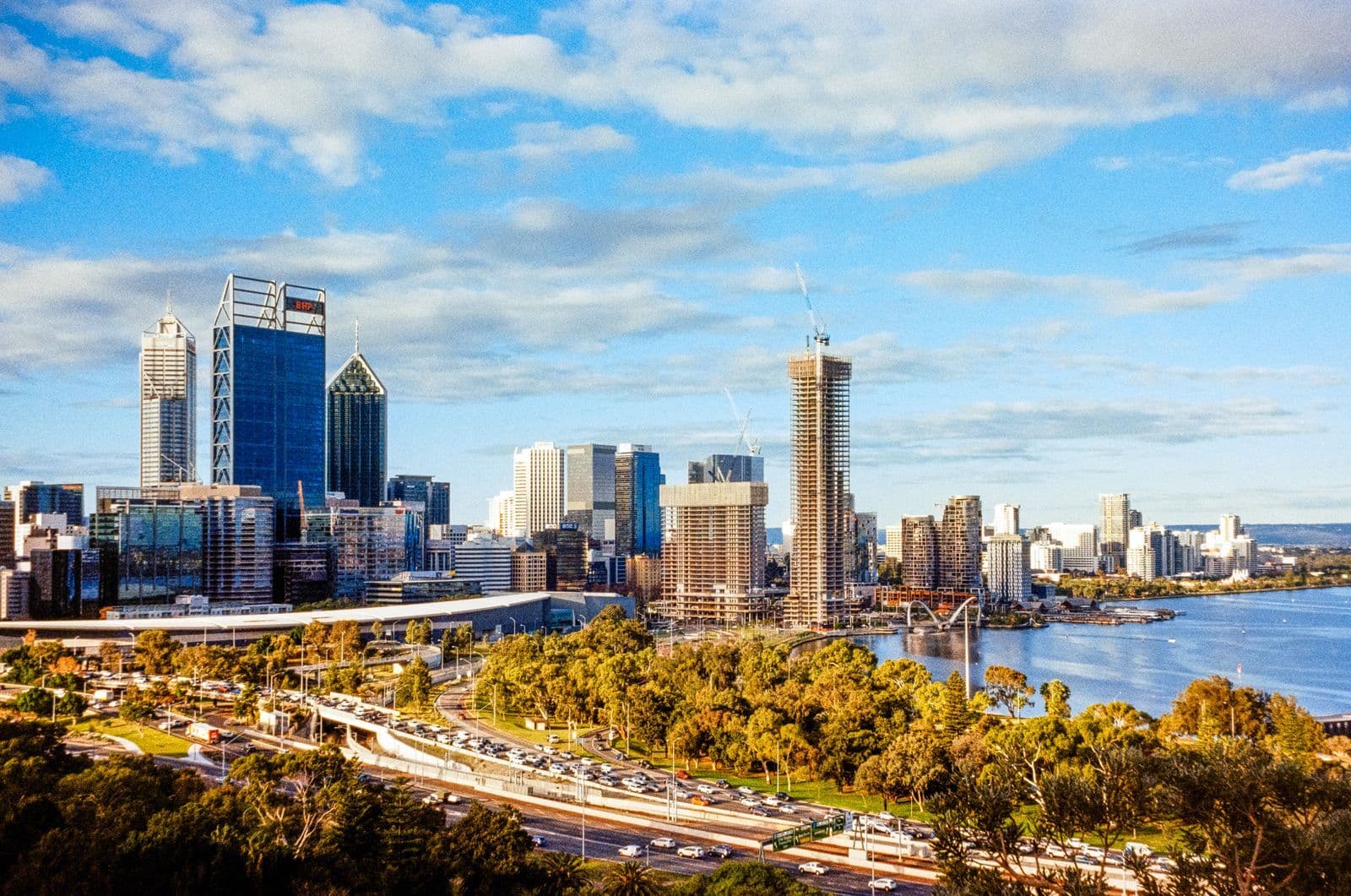 The Perth city skyline beside the Swan River on a sunny day, with traffic streaming along a curving highway in the foreground.