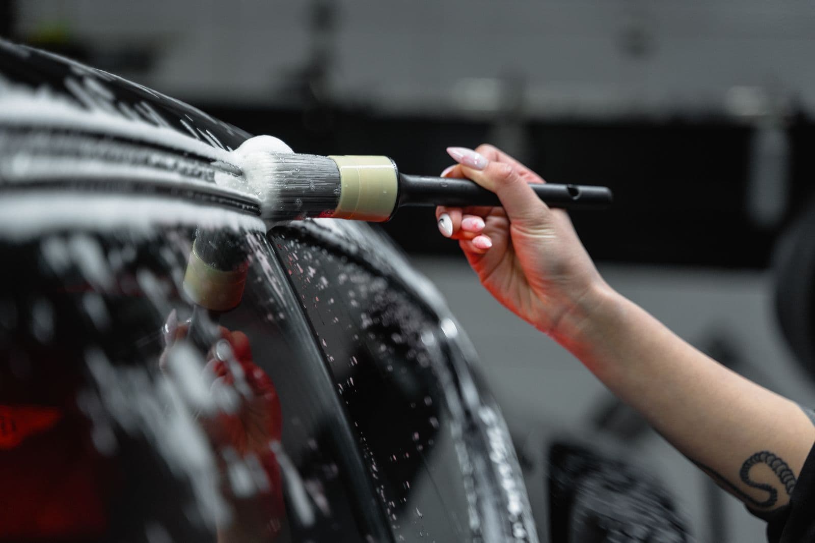 A hand working a soft detailing brush through soap foam along the window trim of a dark car.