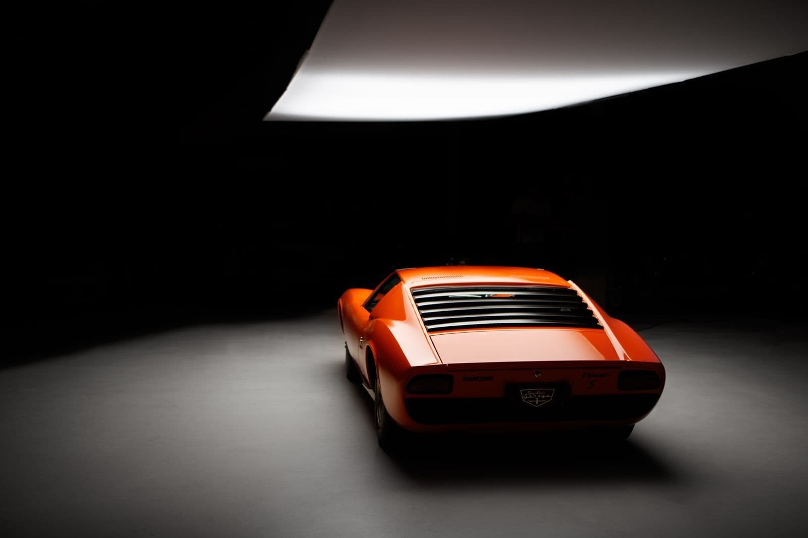 The rear of an orange Lamborghini Miura with its louvred engine cover, lit by a single overhead light in a dark studio.