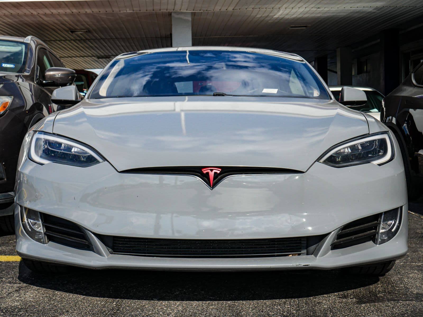 The front of a pale grey Tesla Model S parked under a carport between other cars.