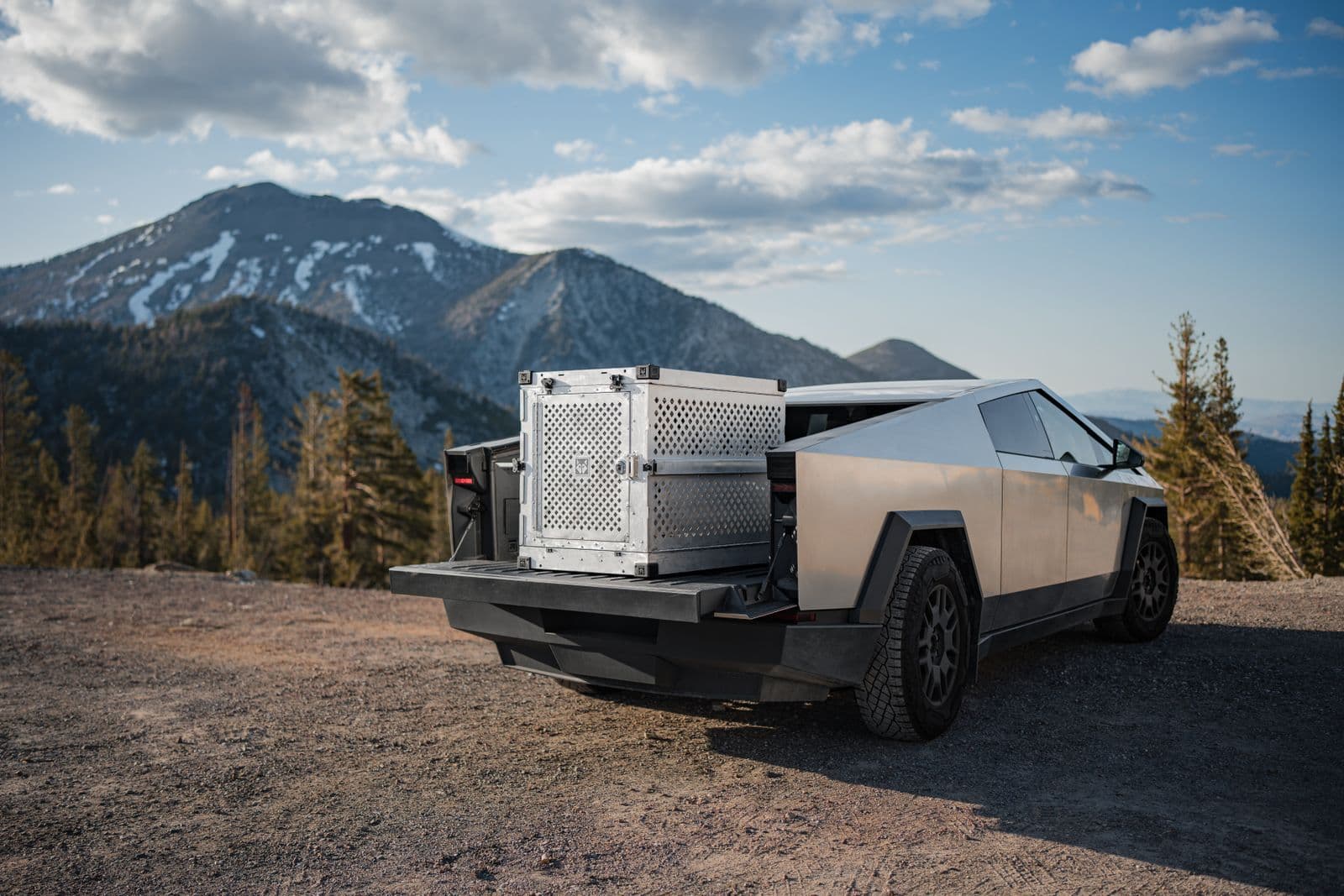 An angular stainless-steel electric pickup parked on gravel with its tailgate down, snow-streaked mountains behind it.