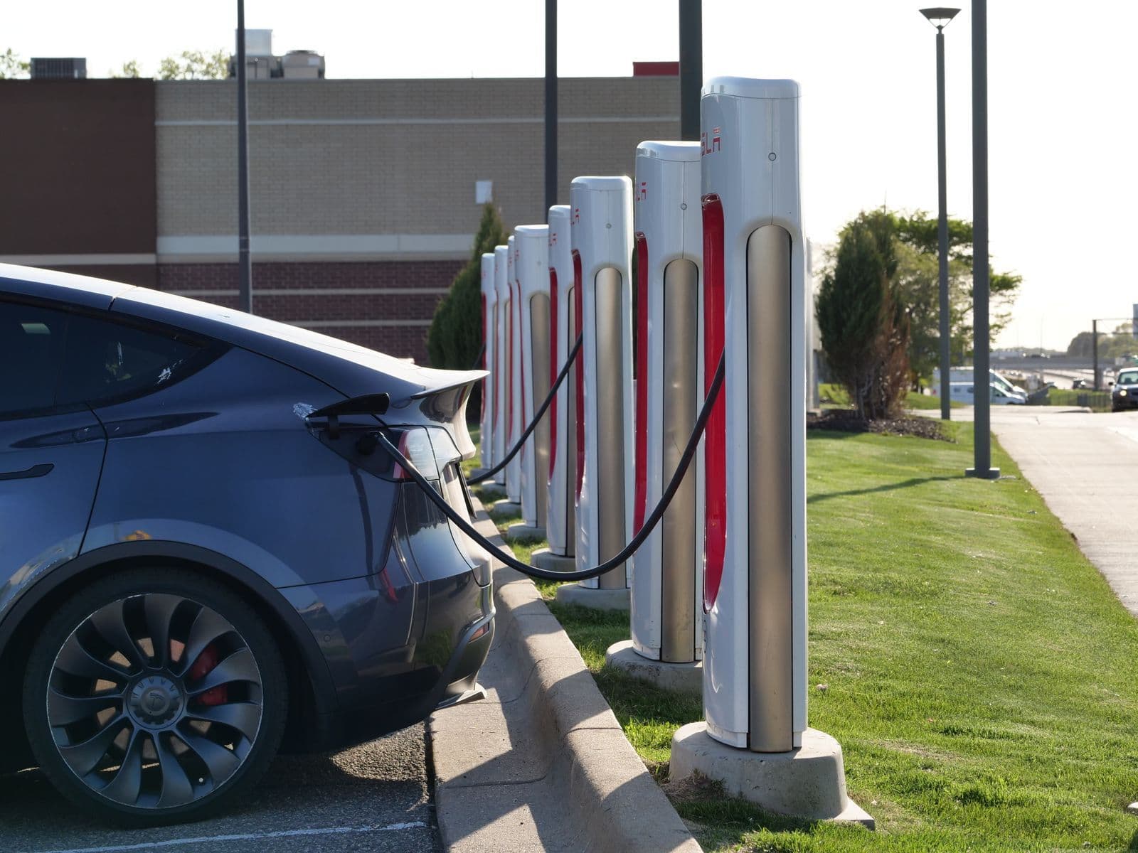 A grey electric car plugged into the first of a long row of fast chargers in a car park.