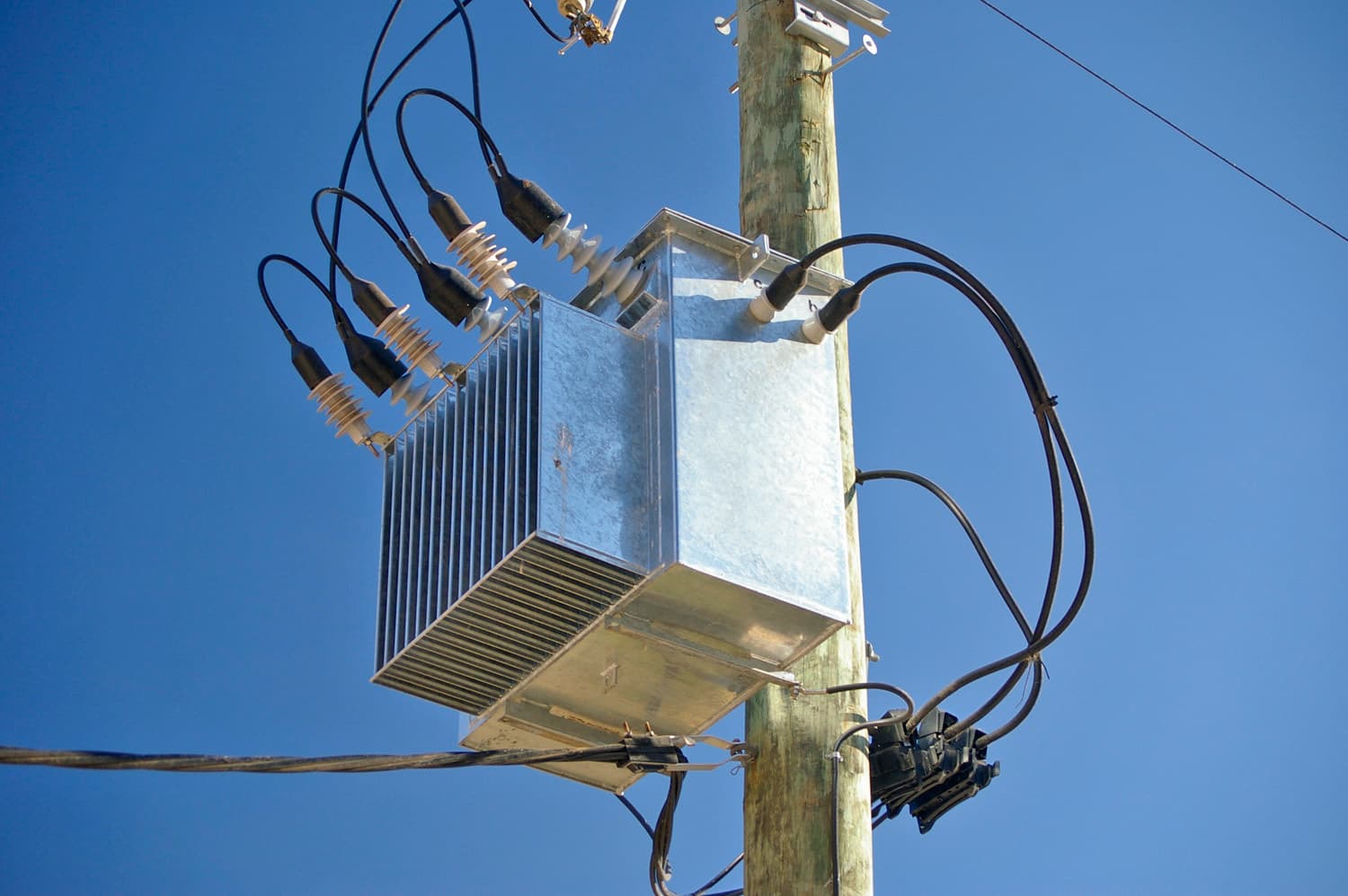 A three-phase pole-mounted electrical distribution transformer bolted to a timber power pole, with overhead lines running off against a pale sky.