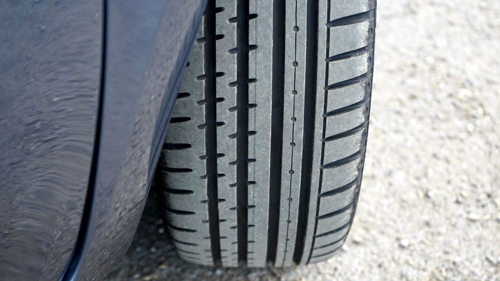 A close-up of a car tyre's tread grooves where the rubber meets a damp gravel road surface.