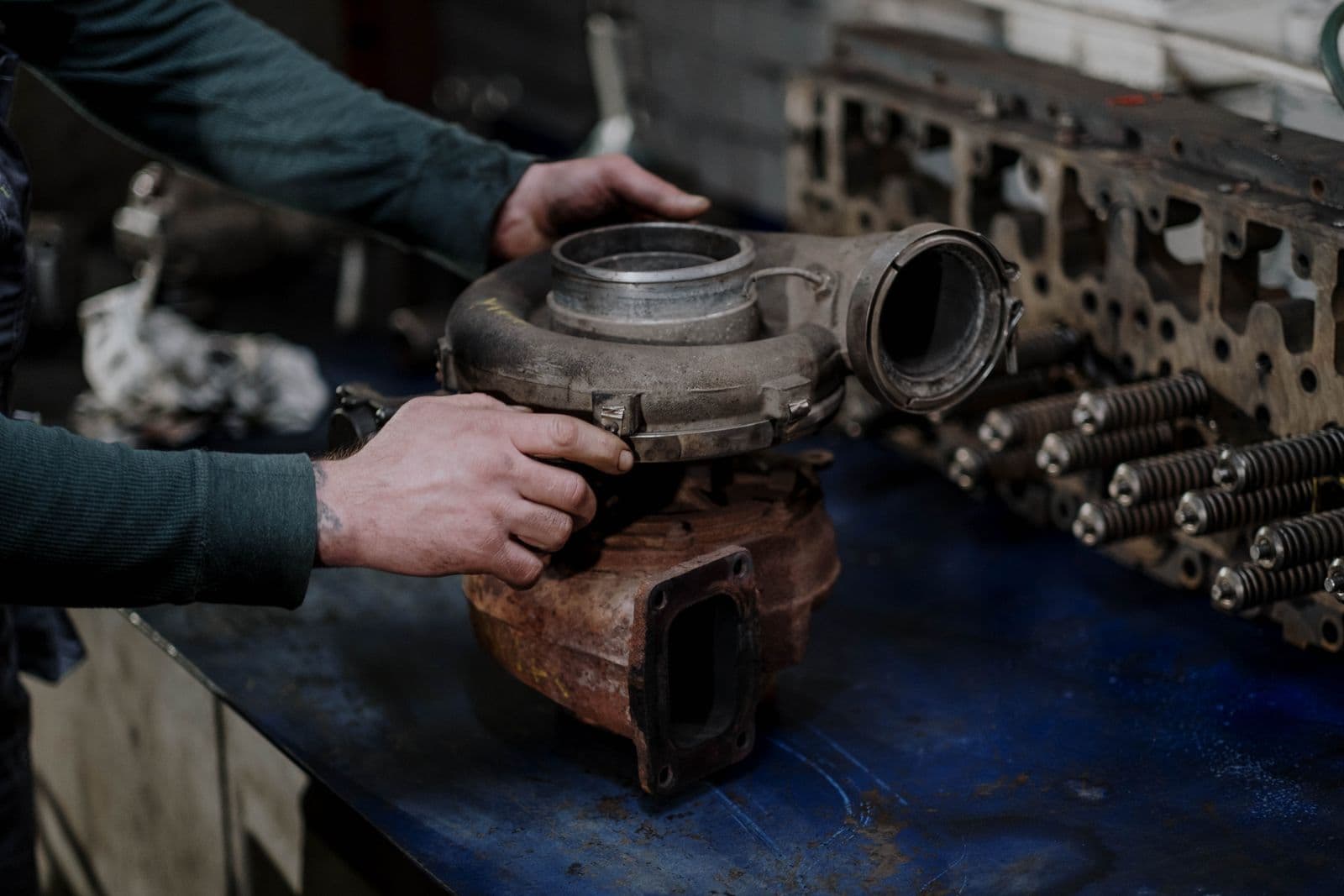 A mechanic lifts a used turbocharger off a workbench beside a stripped cylinder head and its valve springs.