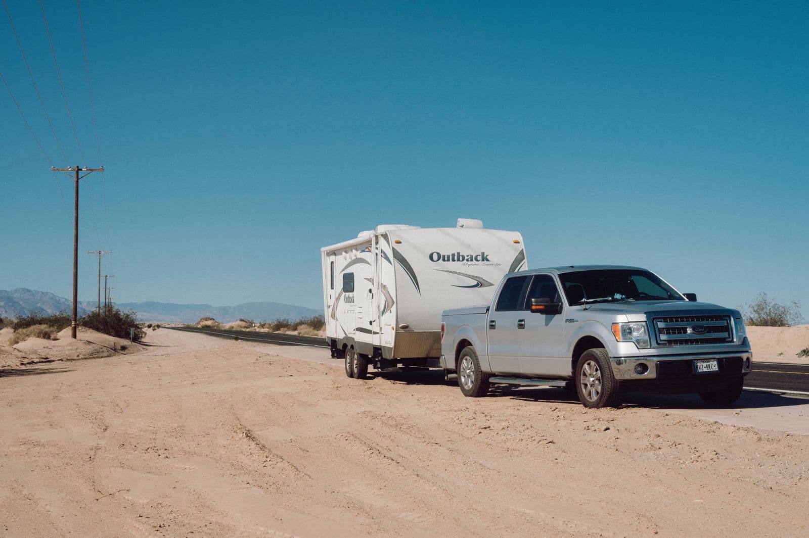 A silver dual-cab ute towing a white caravan along a dusty desert road under a clear sky.