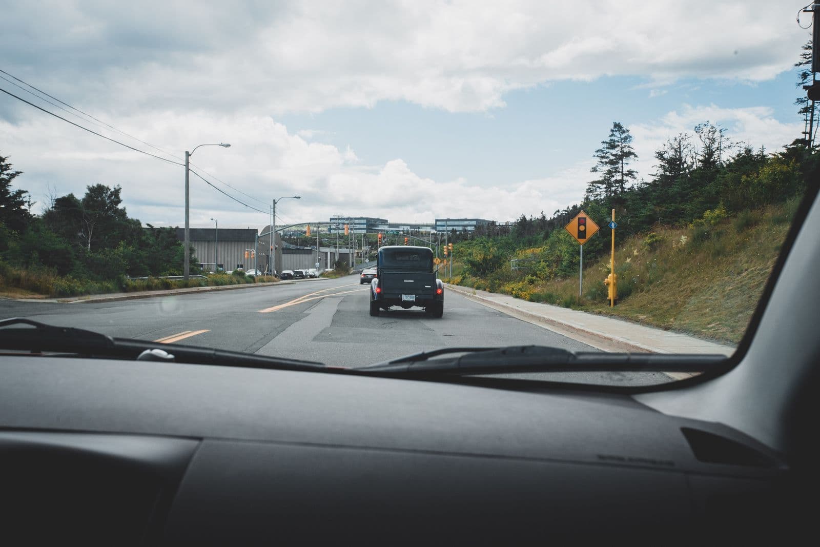 The view through a car's windscreen over the dashboard, following a dark vehicle along a multi-lane road under a cloudy sky.