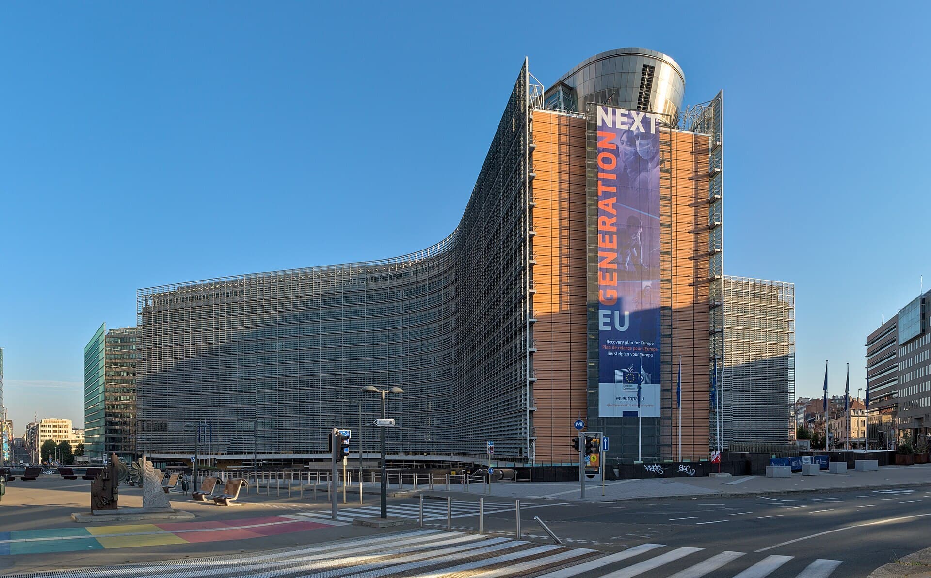 The curved glass-and-steel Berlaymont building in Brussels, headquarters of the European Commission, under a clear blue sky with a pedestrian crossing in front.