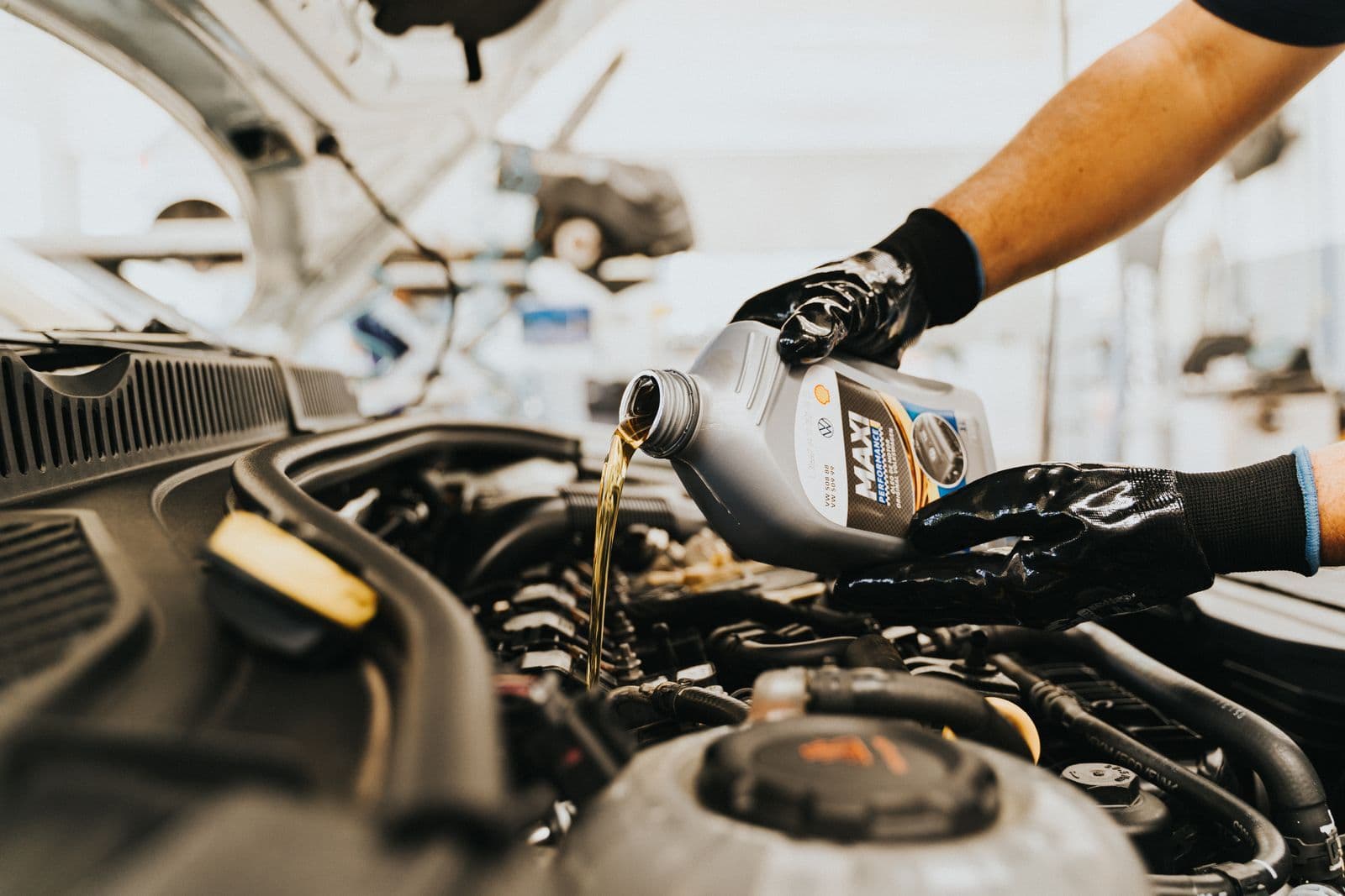 A mechanic in black gloves pours fresh engine oil from a grey bottle into the filler of an open car engine bay.