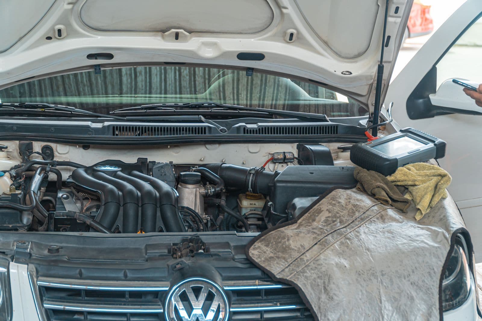 The open bonnet of a white Volkswagen showing the black plastic intake manifold on the engine, with a diagnostic scan tool resting on a cloth over the wing.