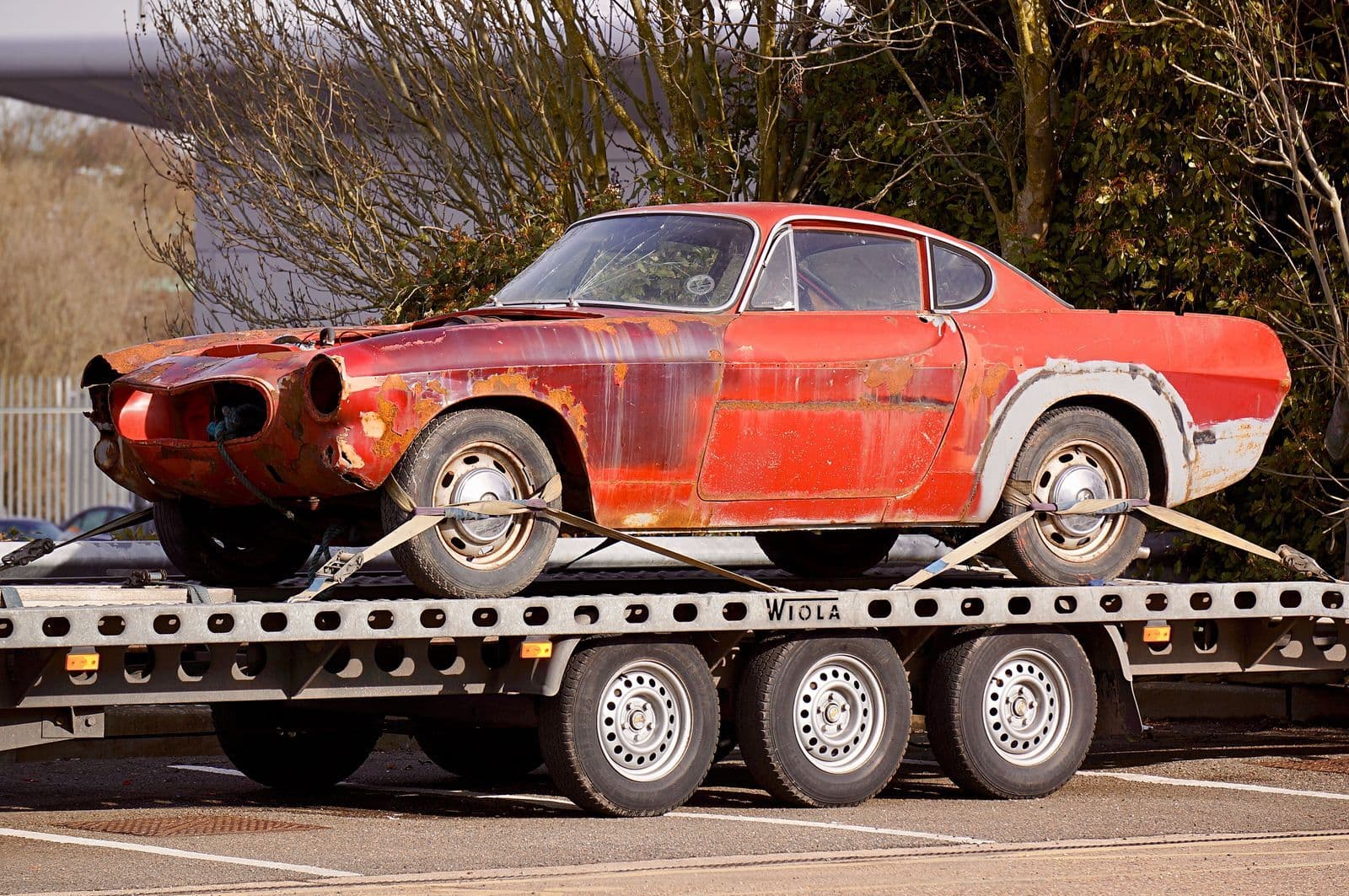 A faded red classic coupe strapped down on the deck of a flatbed car trailer.