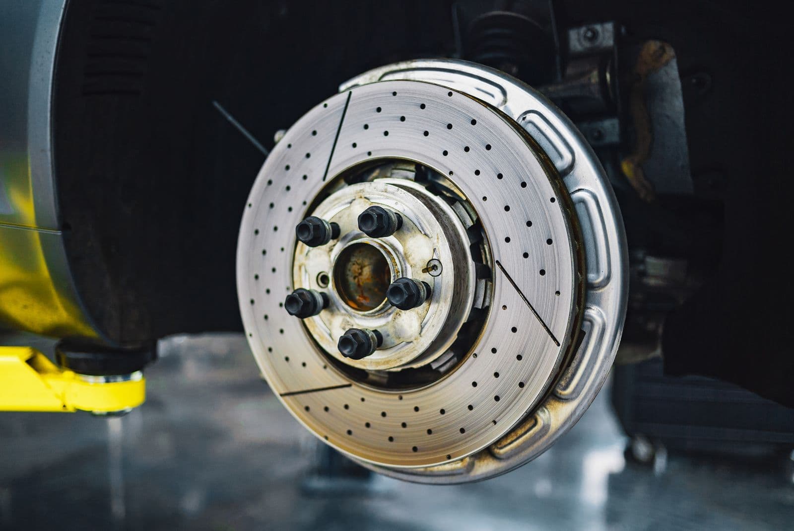 A cross-drilled brake disc and hub on a car with the wheel removed, raised on a workshop hoist.