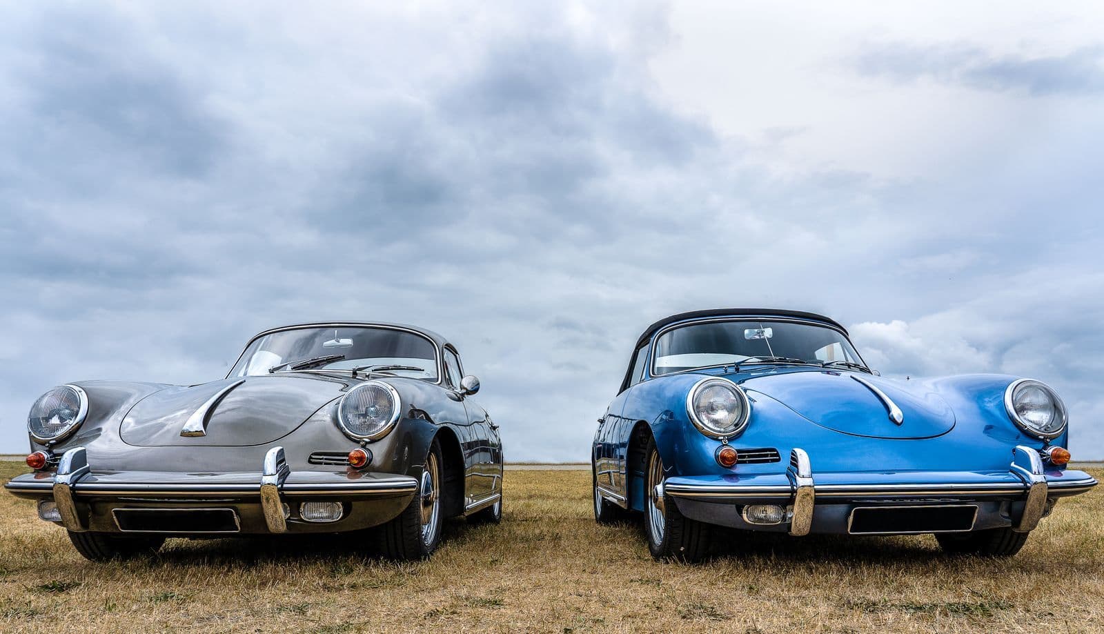 Two classic Porsche 356 sports cars, one silver coupe and one blue cabriolet, parked side by side in a grass field under a heavy grey sky.