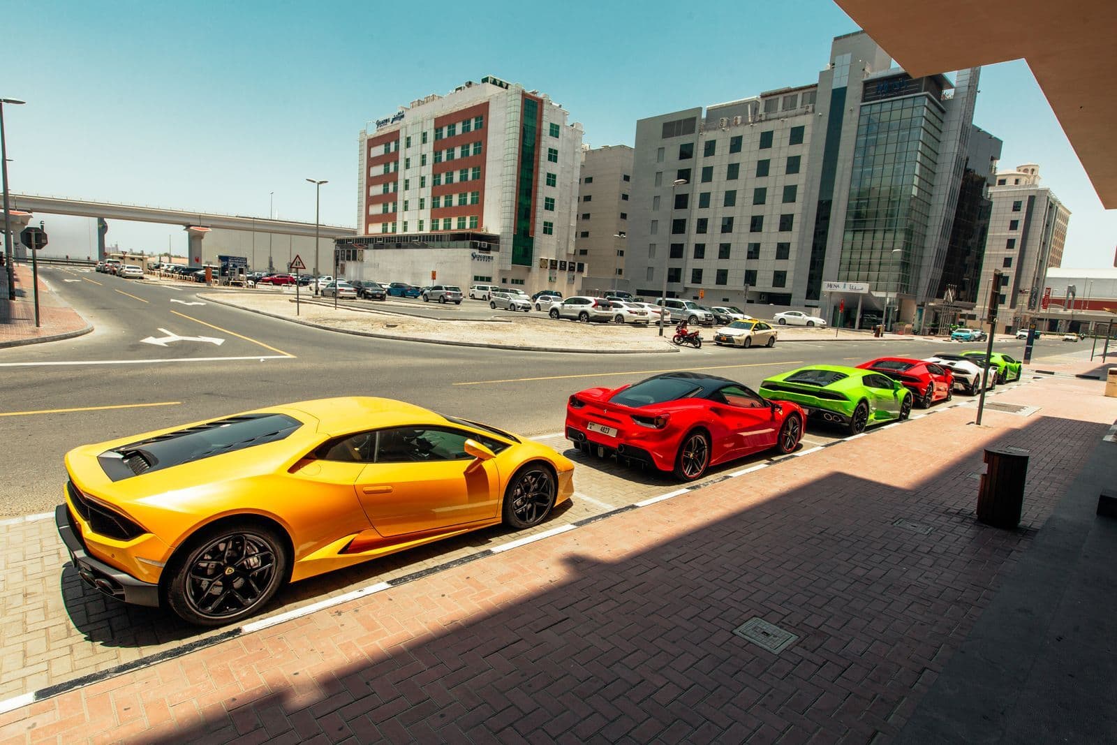 A row of brightly coloured supercars, led by a yellow Lamborghini and a red Ferrari, parked along a kerb in a city street.
