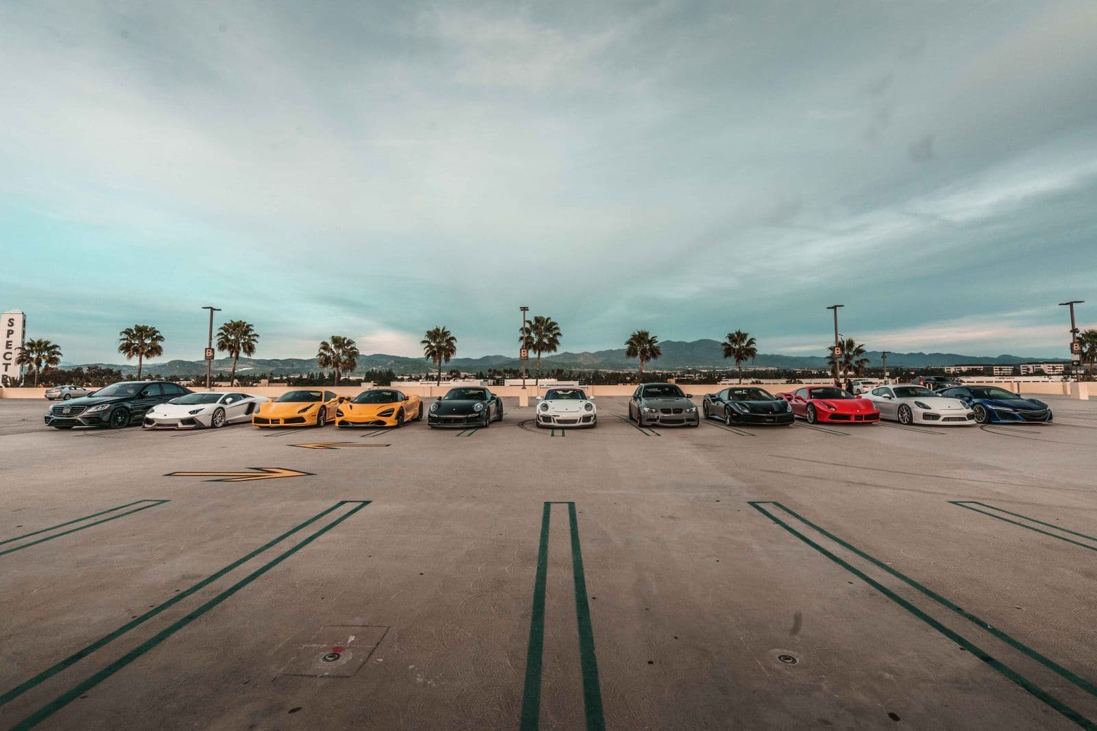 A dozen sports and performance cars parked in a wide fan formation on an open rooftop car park, with palm trees and hills behind.